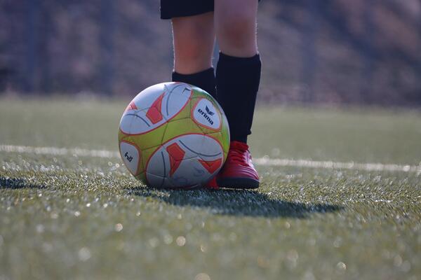 Das Bild zeigt die Beine eines jugendlichen Fußballers mit Fußballschuhen und Stulpen und einen bunten Fußball auf grünem Rasen