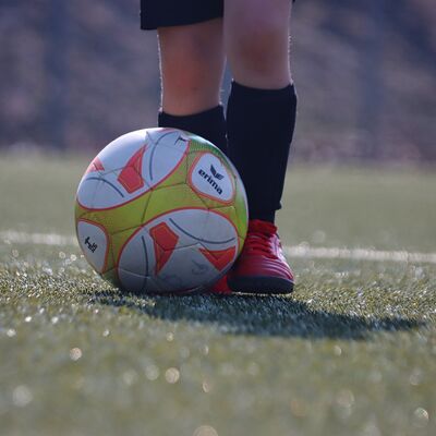 Das Bild zeigt die Beine eines jugendlichen Fußballers mit Fußballschuhen und Stulpen und einen bunten Fußball auf grünem Rasen