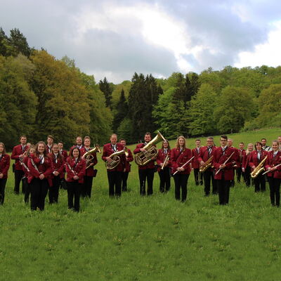 Das Foto zeigt Musiker des Musikzugs Bottendorf in rot-schwarzer Uniform aufgestellt mit ihren Instrumenten. Die Musiker stehen auf einer grünen Wiese im Hintergrund sind Bäume. 